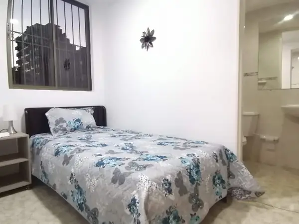 Habitación con cama, mesa de noche y ventana; a un lado se ve un baño